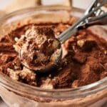 A close-up of a glass bowl filled with tiramisu ice cream, topped with cocoa powder, as an ice cream scoop lifts a creamy portion.