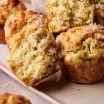 Close-up of golden-brown muffins, one cut in half to show a moist, crumbly interior with visible shreds of zucchini.
