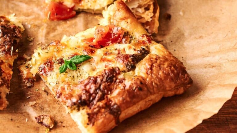 A slice of cheesy tomato tart with a golden, flaky crust sits on brown parchment paper, garnished with a small basil leaf.