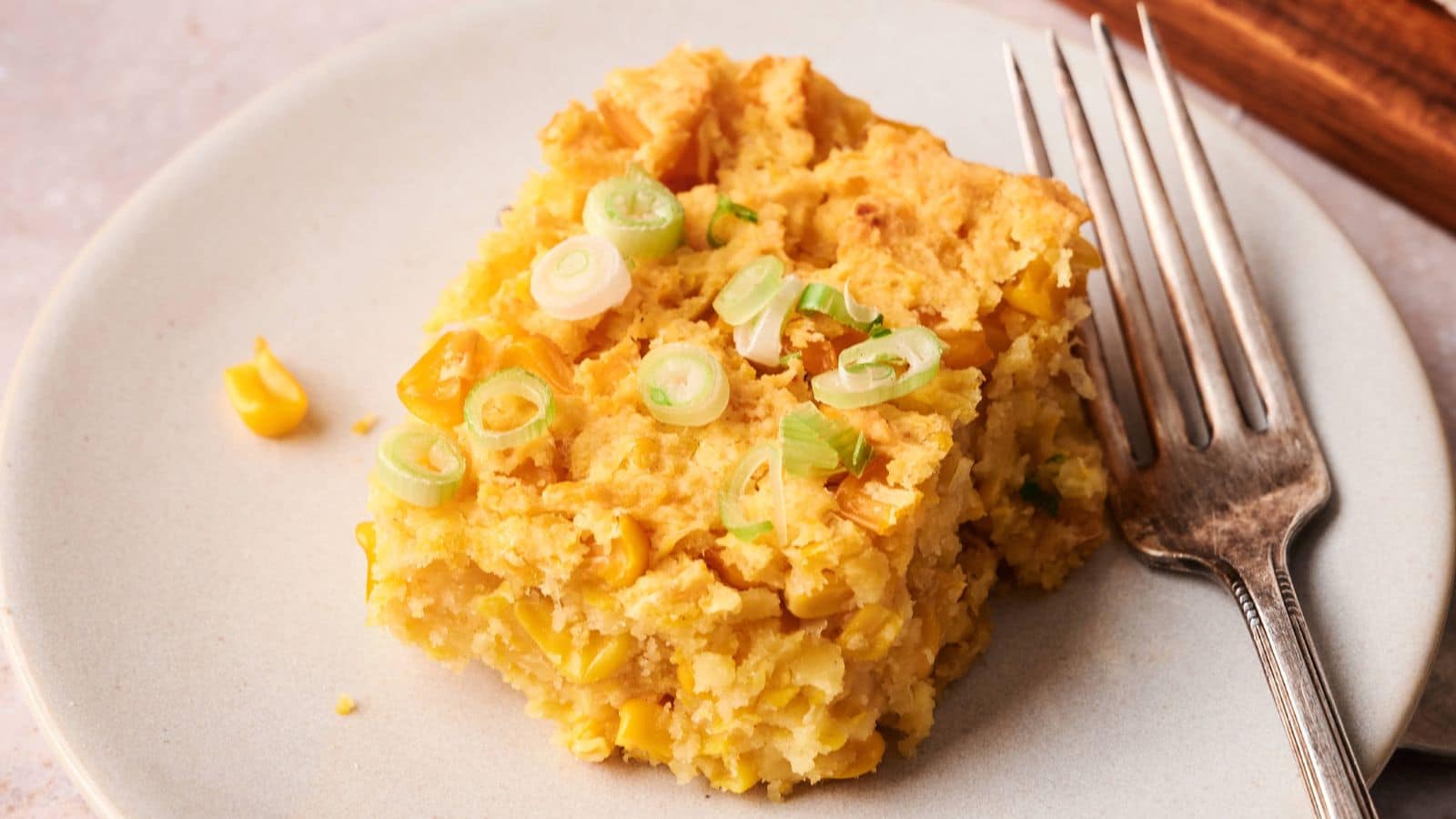 A square piece of cornbread with corn kernels and sliced green onions on top sits on a white plate next to a fork.