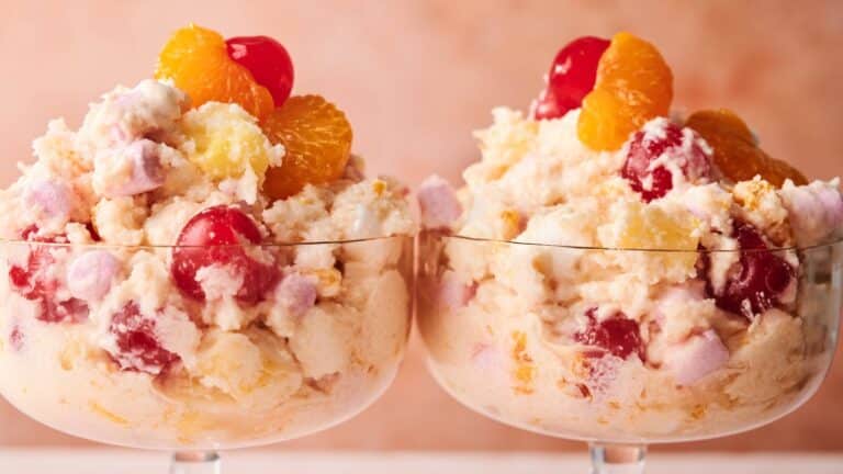Two glass dessert bowls filled with ambrosia salad, featuring whipped cream, marshmallows, cherries, pineapple, and mandarin orange segments against a light pink background.