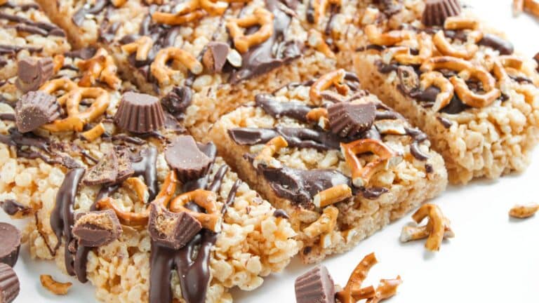 Rice crispy treats topped with pretzel pieces, mini peanut butter cups, and drizzled with chocolate, cut into squares and arranged on a white surface.