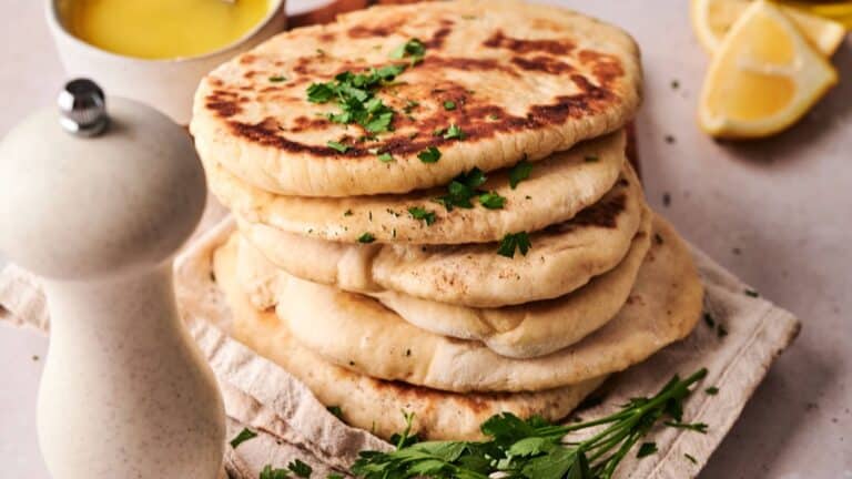 A stack of flatbreads garnished with chopped herbs sits on a cloth, with a pepper grinder, lemon wedges, a bowl of oil, and fresh parsley nearby.