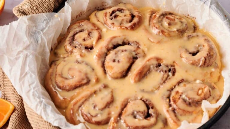 A baking dish lined with parchment paper holds several iced cinnamon rolls covered in glaze.