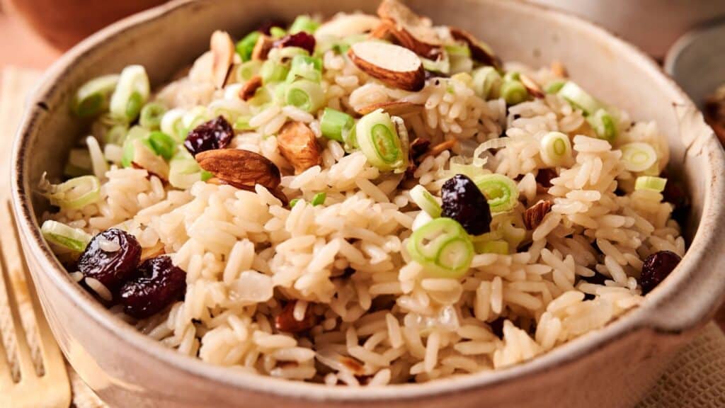 A bowl of cooked rice topped with sliced green onions, almonds, and dried cranberries.