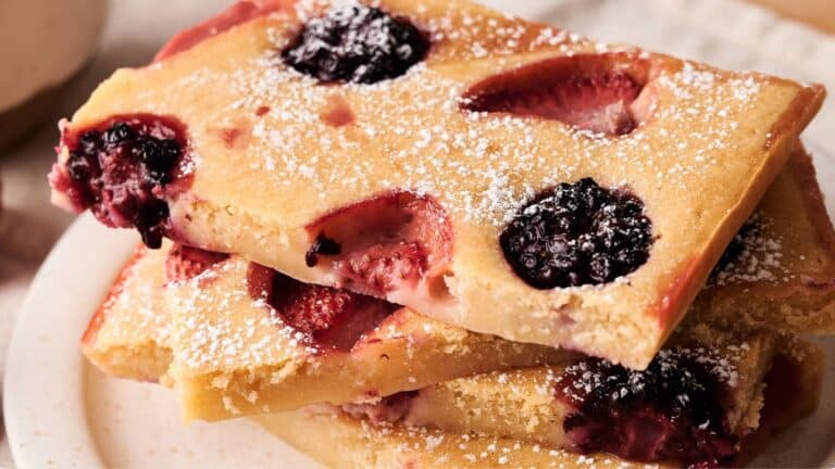 Three rectangular pieces of baked pancake topped with powdered sugar, featuring baked-in blackberries and strawberries, stacked on a white plate.