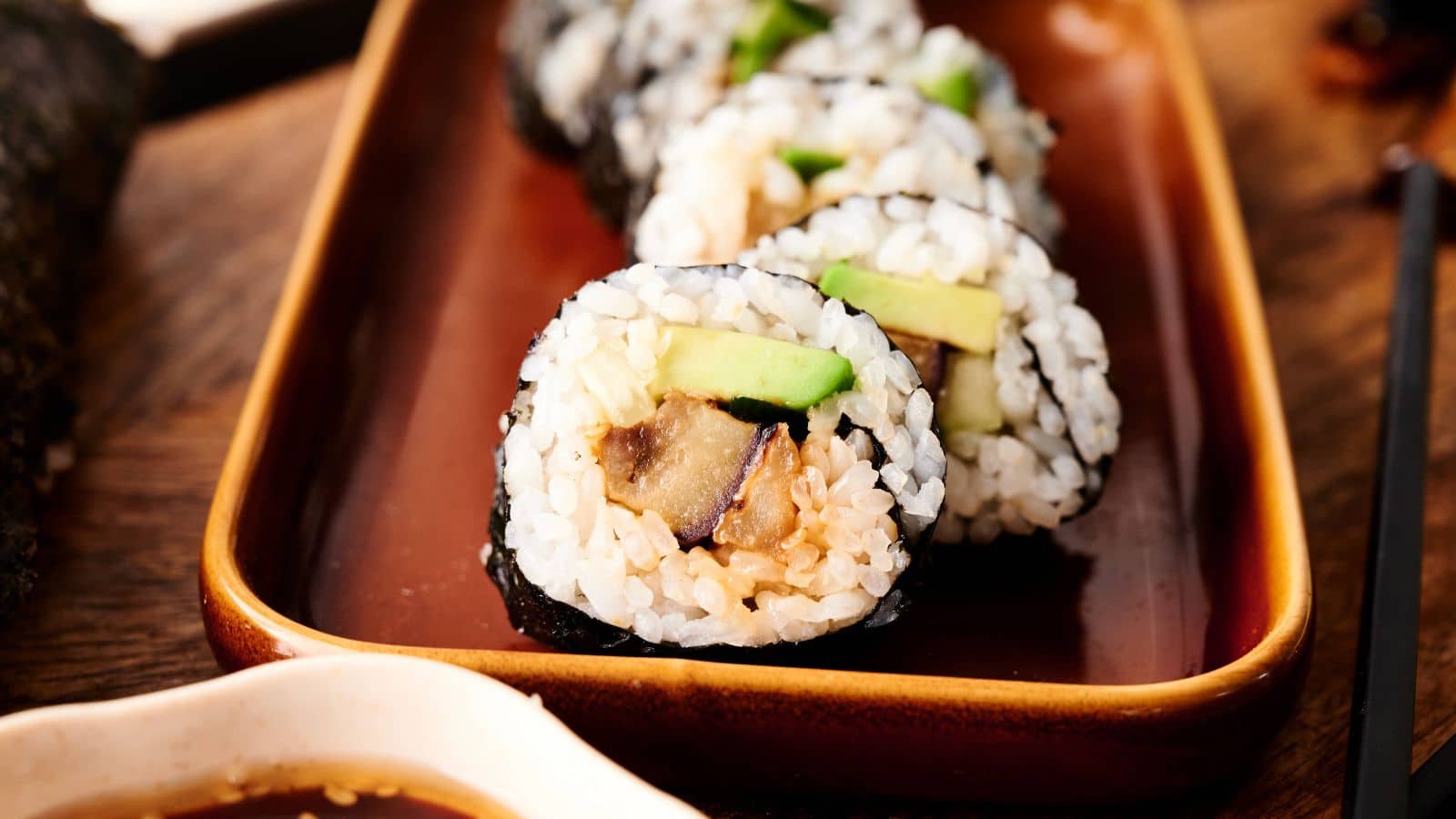 Close-up of three sushi rolls filled with rice, avocado, and vegetables, served on a brown rectangular plate with a small dish of soy sauce nearby.
