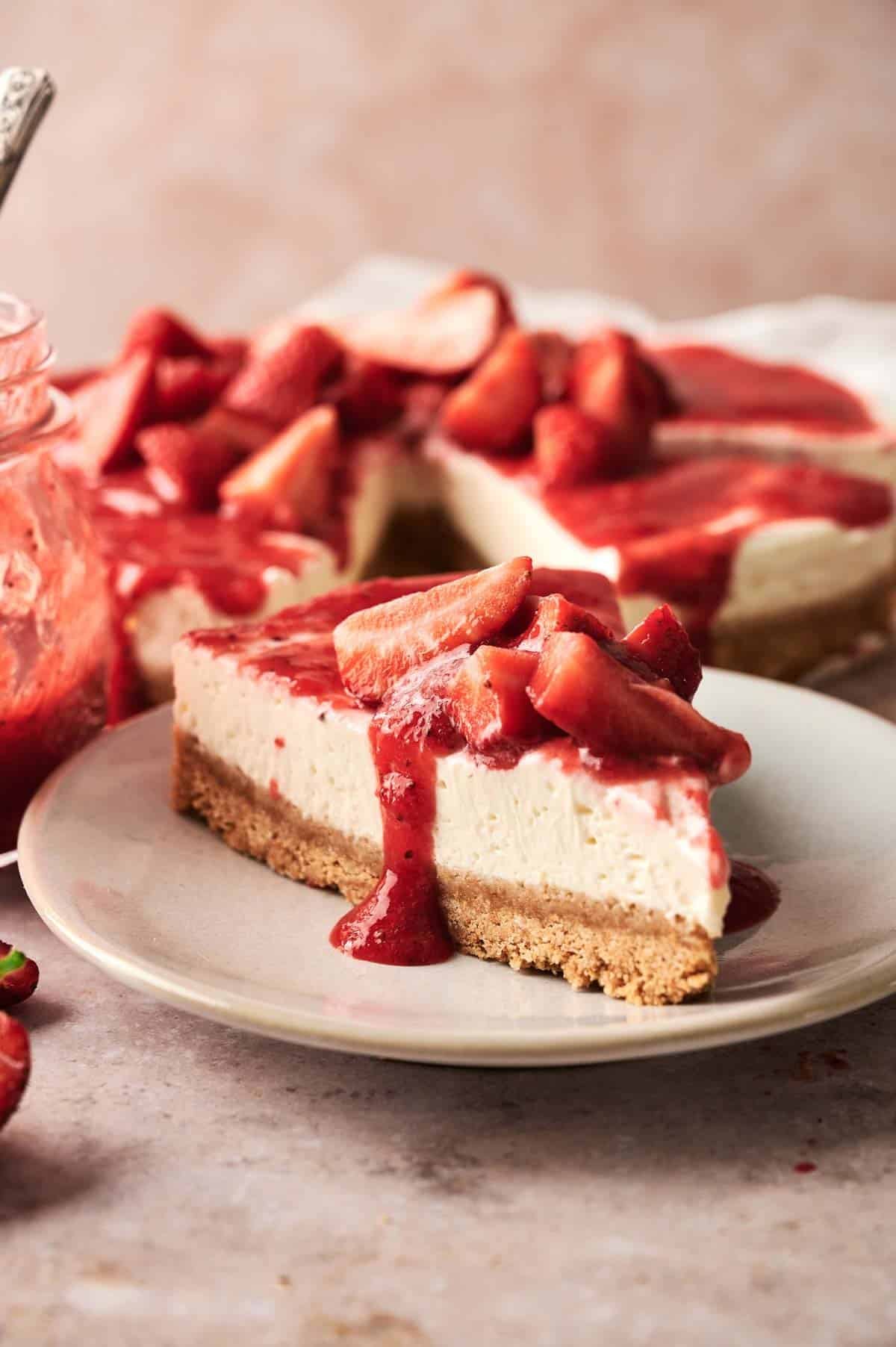A slice of no bake strawberry cheesecake topped with strawberry sauce and fresh strawberries sits on a plate; the remaining cheesecake is in the background.