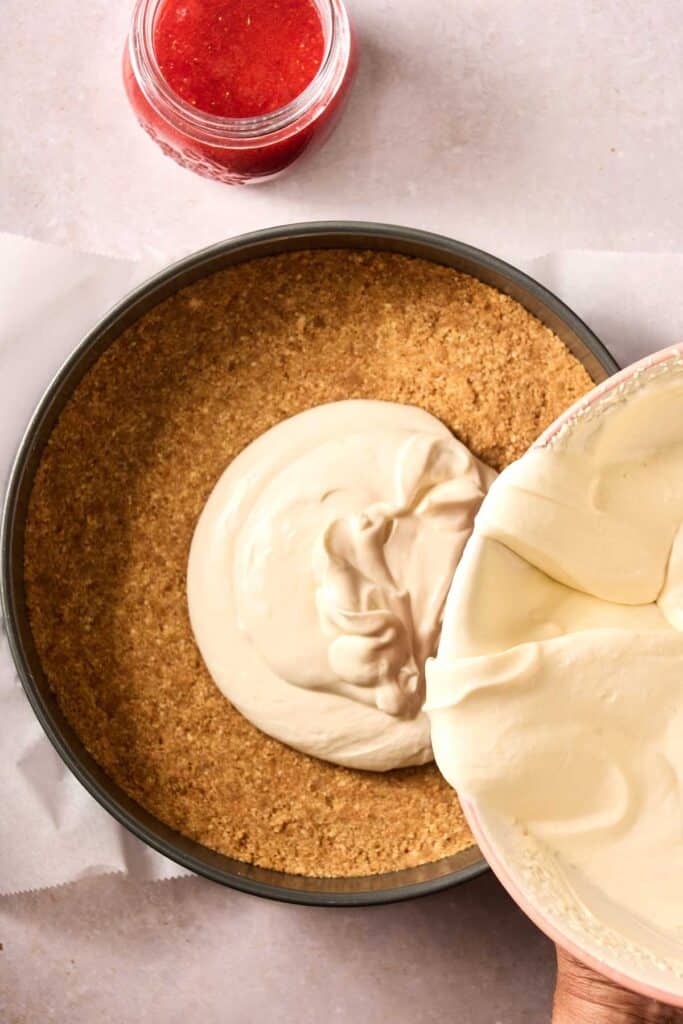 Cheesecake filling being poured into a prepared graham cracker crust in a round pan, with a jar of red sauce nearby—perfect for making a no bake strawberry cheesecake on a light surface.