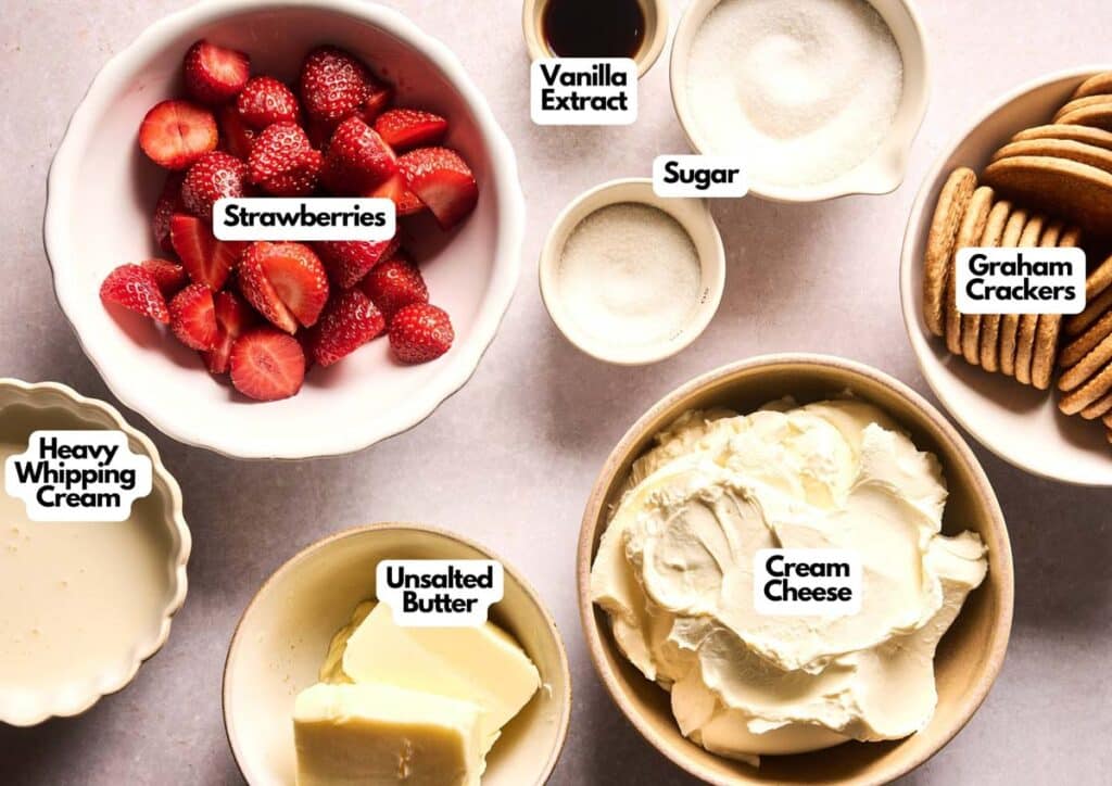 Top-down view of labeled bowls with ingredients for a no bake strawberry cheesecake: strawberries, sugar, vanilla extract, graham crackers, cream cheese, unsalted butter, and heavy whipping cream.