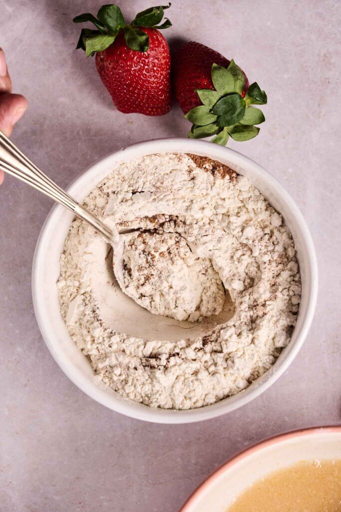 A hand stirs flour in a white bowl with a metal spoon, preparing strawberry bread. Two strawberries are placed on the surface next to the bowl.