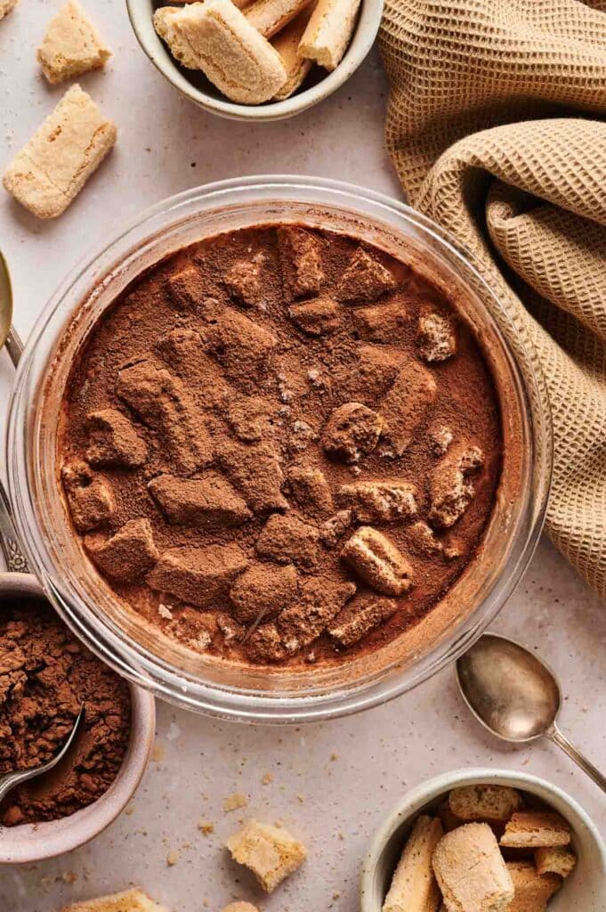 A glass dish filled with tiramisu ice cream, topped with cocoa powder and ladyfinger pieces, surrounded by bowls of ingredients and a beige cloth.