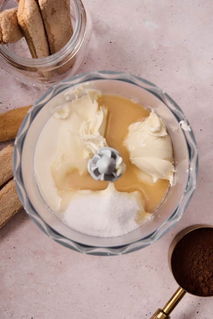 A food processor bowl containing mascarpone cheese, sugar, and liquid ingredients for tiramisu ice cream, surrounded by ladyfinger biscuits and a measuring cup of cocoa powder.