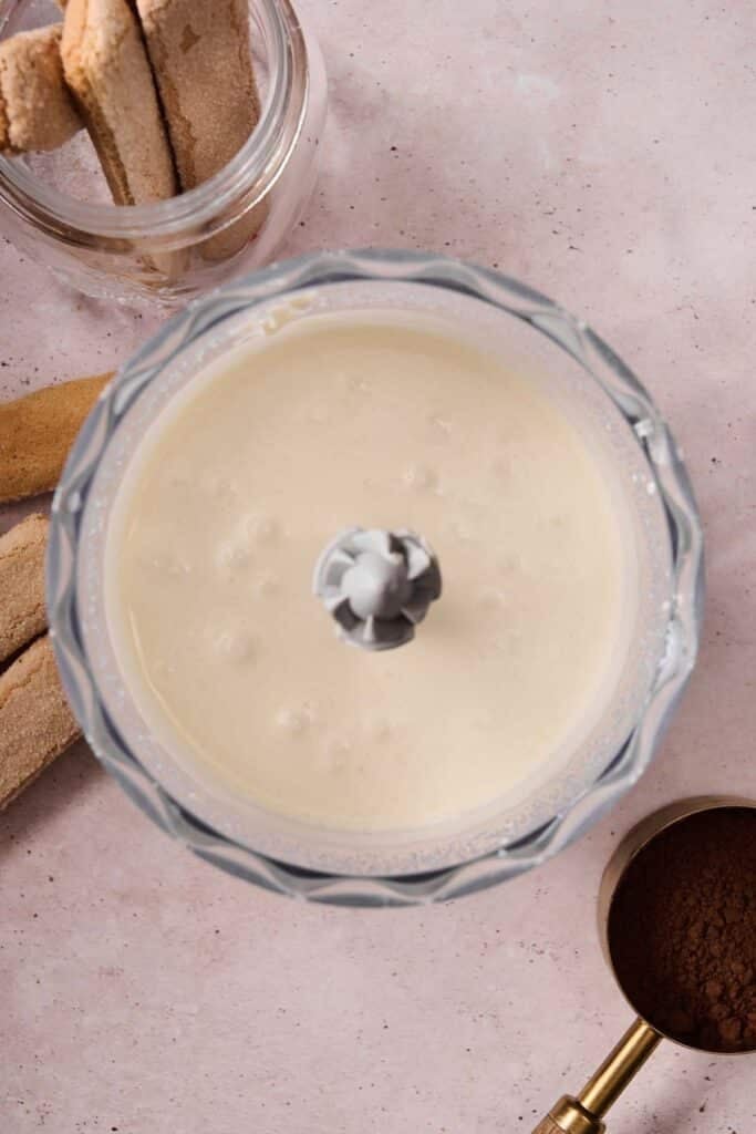 A food processor bowl with smooth tiramisu ice cream mixture, surrounded by ladyfingers in a jar, loose ladyfingers, and a measuring cup of cocoa powder on a light pink surface.