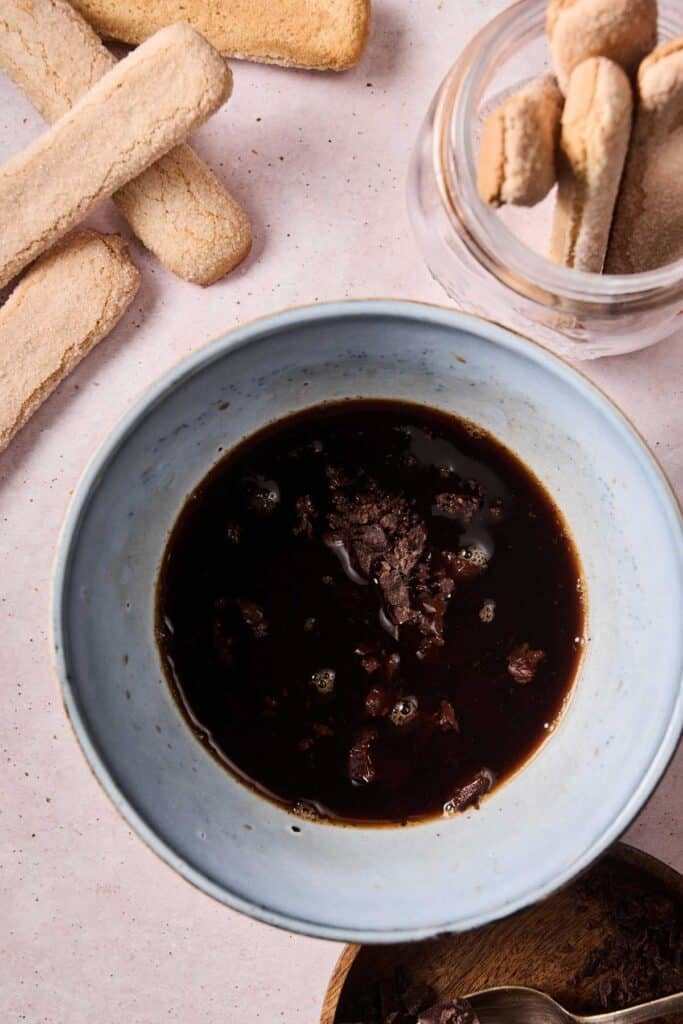 A blue bowl containing dark liquid with chocolate pieces, surrounded by ladyfingers and a jar with more ladyfingers on a light surface—perfect ingredients for classic tiramisu ice cream.