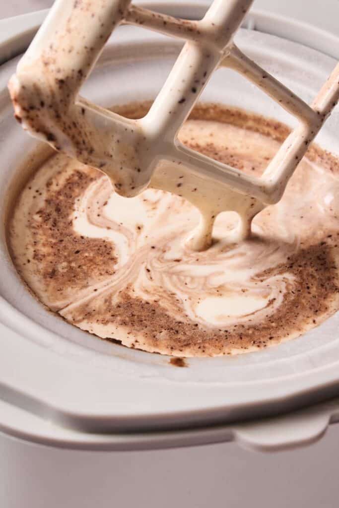 Close-up of an ice cream maker paddle mixing a creamy, swirled tiramisu ice cream with visible chocolate flecks inside the bowl.