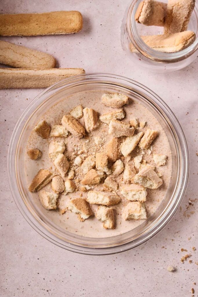 A glass bowl filled with crushed ladyfinger cookies, perfect for making tiramisu ice cream, sits on a light surface, with whole ladyfingers and a jar of more cookies nearby.
