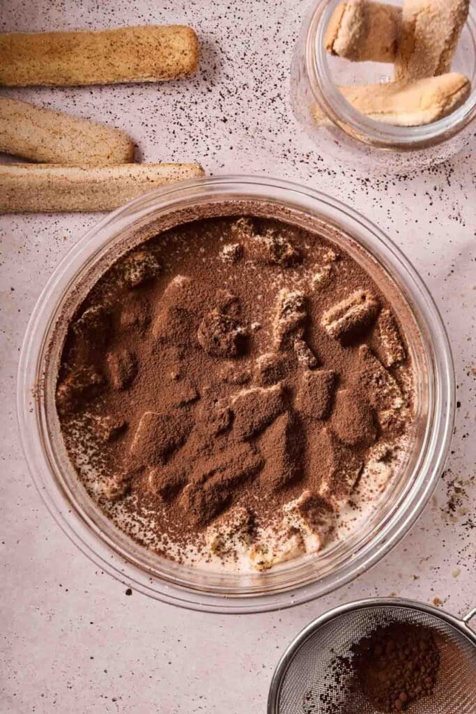 A bowl of tiramisu ice cream topped with cocoa powder sits on a light surface, surrounded by ladyfingers, a jar of more ladyfingers, and a metal sieve.
