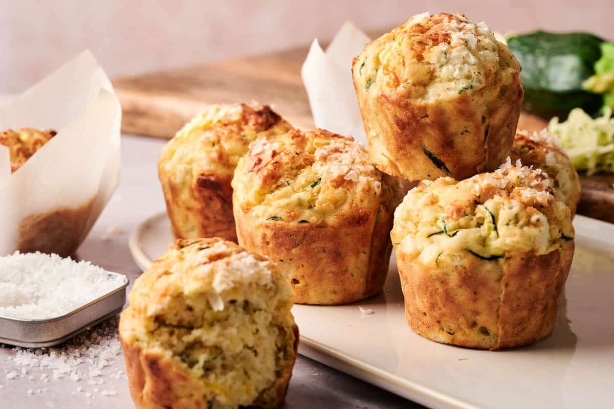 Six savory zucchini muffins are arranged on a plate, topped with grated cheese, with a small dish of shredded cheese nearby and a wooden board in the background.