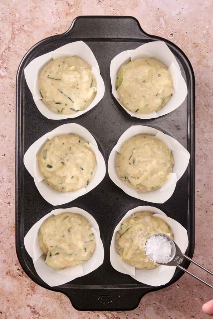 A muffin tin with six paper liners filled with batter, and a hand holding a spoon with salt above one of the muffins.