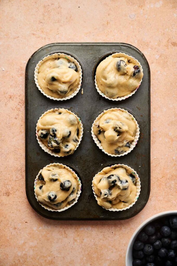 A muffin tin with six unbaked Blueberry Muffins batter portions in white paper liners rests on a light brown surface beside a bowl of fresh blueberries.