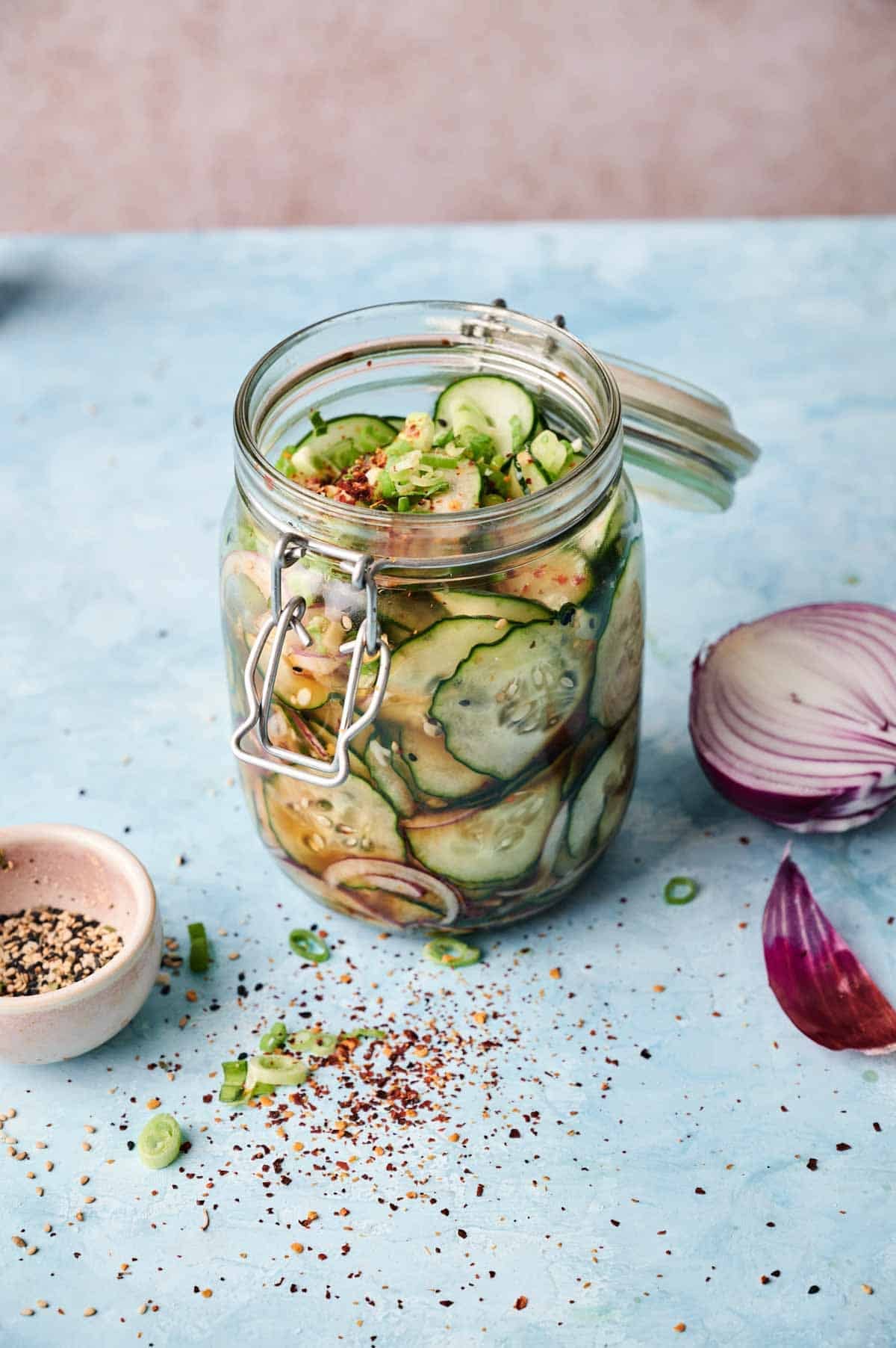 A Cucumber Salad Jar filled with sliced cucumbers and red onions in pickling liquid sits on a light blue surface next to spices and a bowl of pepper.