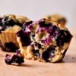 A close-up of a Blueberry Muffin with a bite taken out, showing the moist interior and whole blueberries. Other blueberry muffins and scattered berries are blurred in the background.