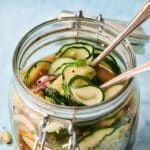 A Cucumber Salad Jar filled with sliced pickled cucumbers and onions in brine, two metal forks inside, set against a light blue background.