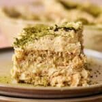A close-up of a Matcha Tiramisu slice dusted with vibrant green matcha powder on a ceramic plate, with more tiramisu in the background.