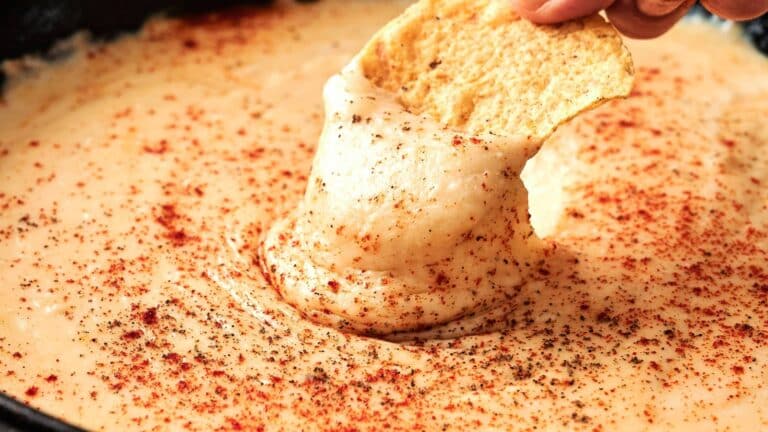 A tortilla chip being dipped into creamy, melted cheese sauce sprinkled with paprika and black pepper.