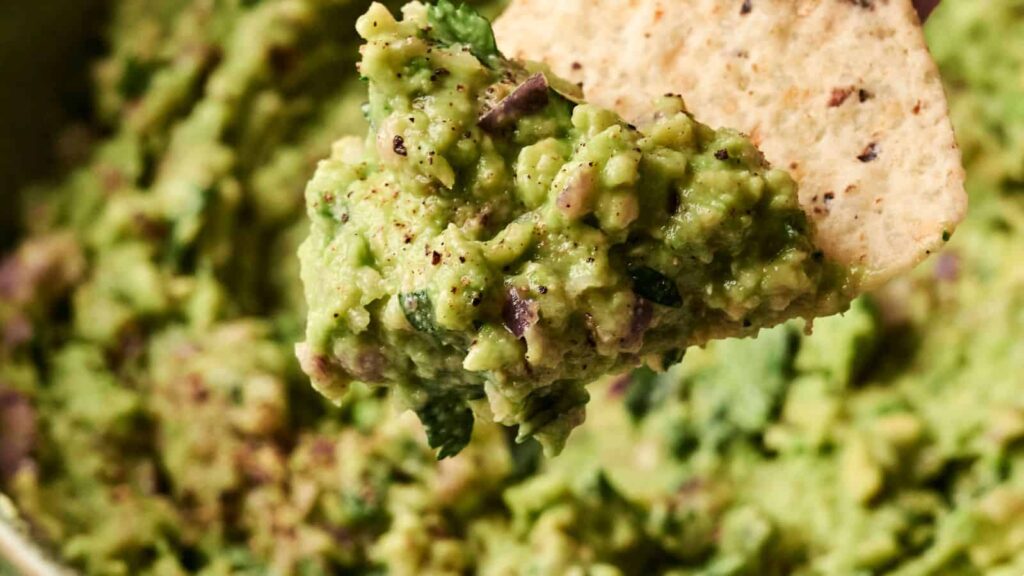 A tortilla chip dipping into a bowl of guacamole with visible pieces of cilantro and seasoning.