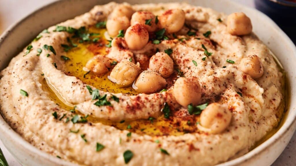 A bowl of hummus topped with whole chickpeas, chopped parsley, paprika, and a drizzle of olive oil.
