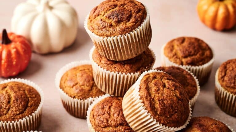 A stack of pumpkin muffins in paper liners on a light surface, with decorative white and orange pumpkins in the background.