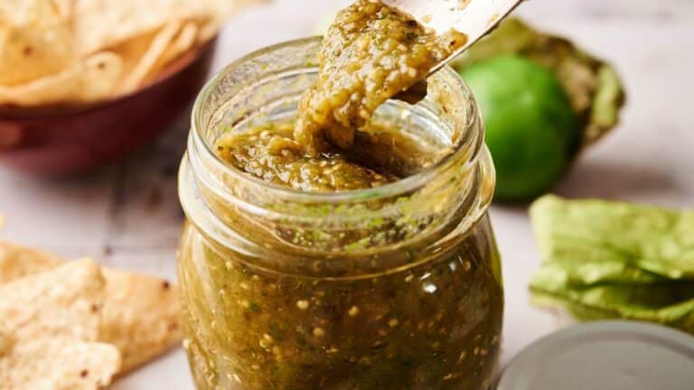 A jar of green salsa with a spoon scooping some out, surrounded by tortilla chips and a fresh tomatillo.