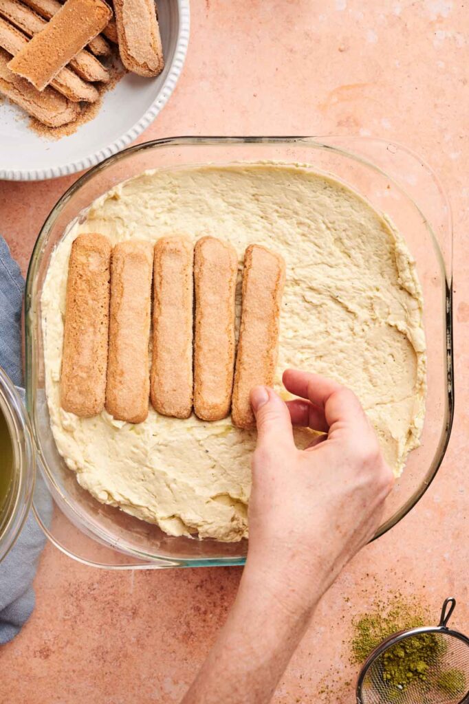 A hand places ladyfingers on a layer of cream in a glass baking dish, preparing Matcha Tiramisu. A bowl of ladyfingers and a sifter with green matcha powder are nearby.