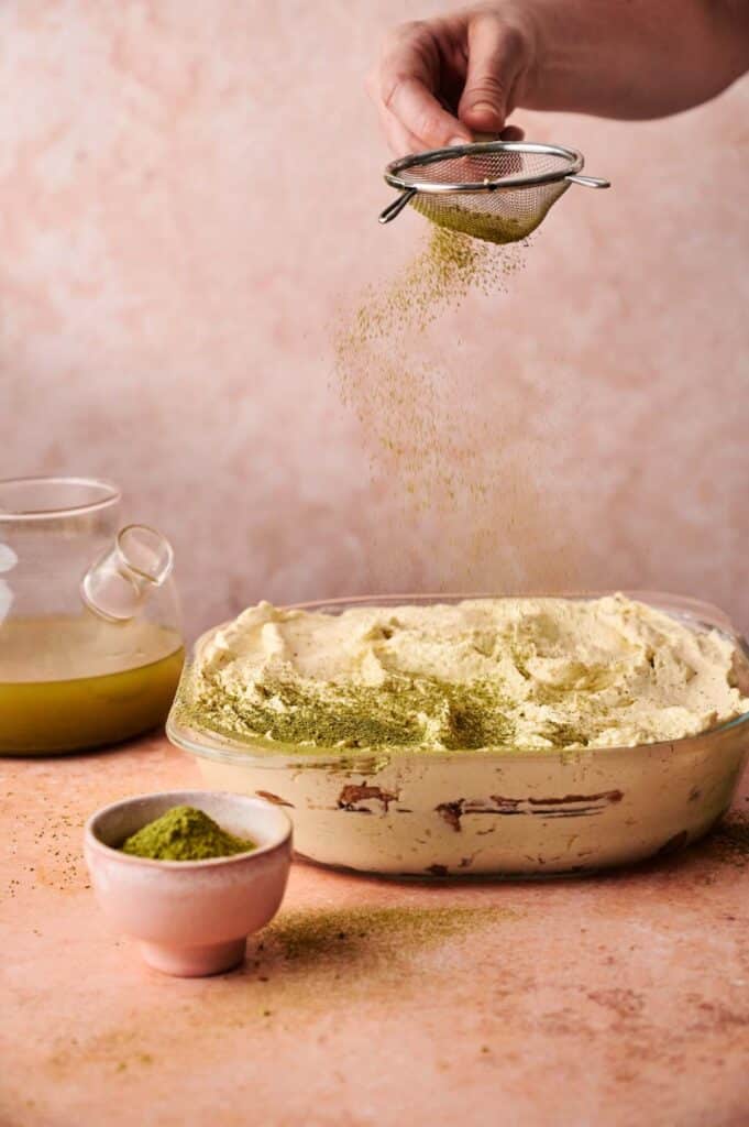 A hand sifts green matcha powder over a dish of creamy Matcha Tiramisu, with a teapot and a small bowl of matcha powder nearby on a light pink surface.
