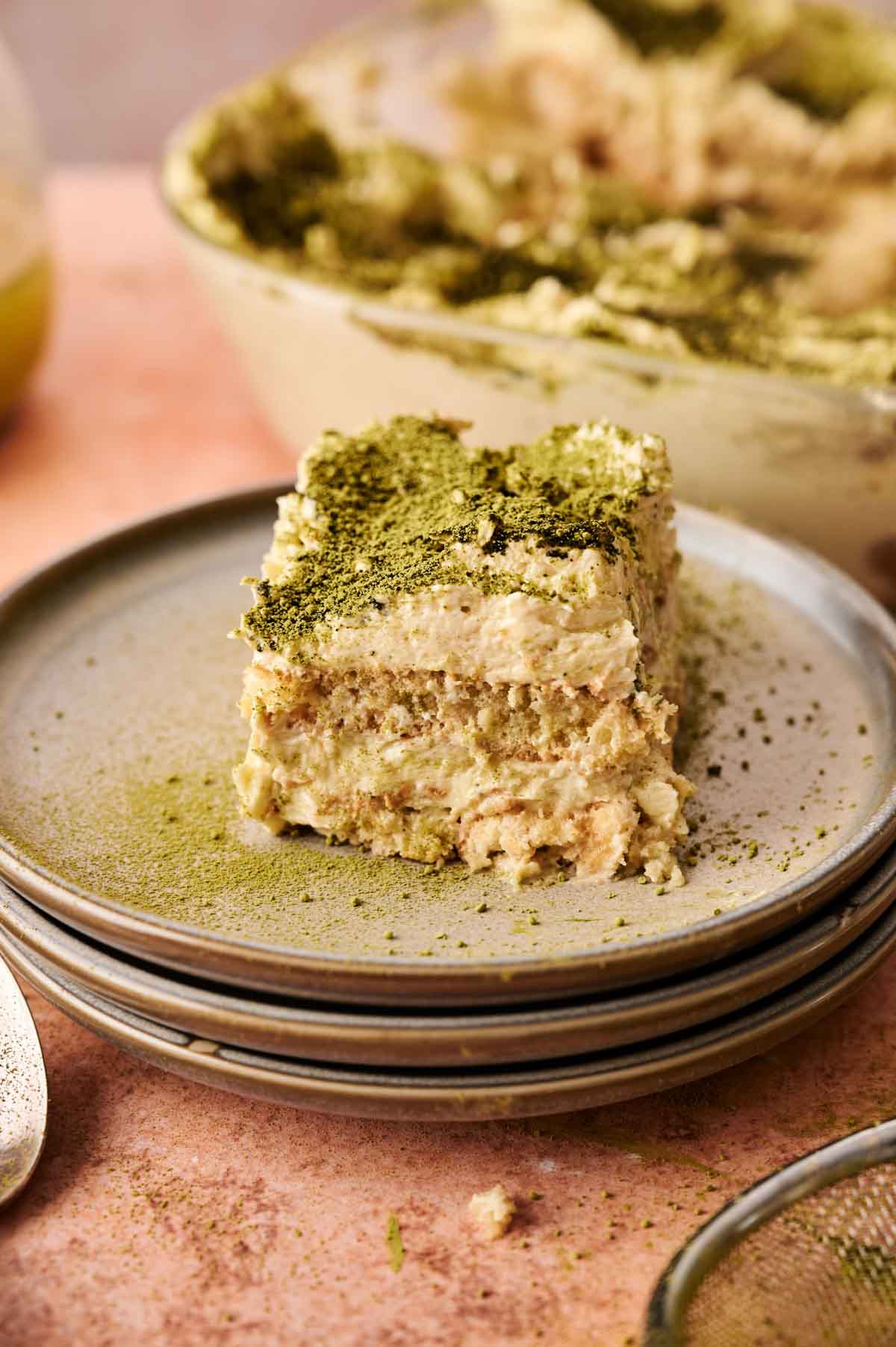A slice of Matcha Tiramisu, dusted with vibrant green matcha powder, rests on a stack of plates while the remaining dessert awaits in the background.