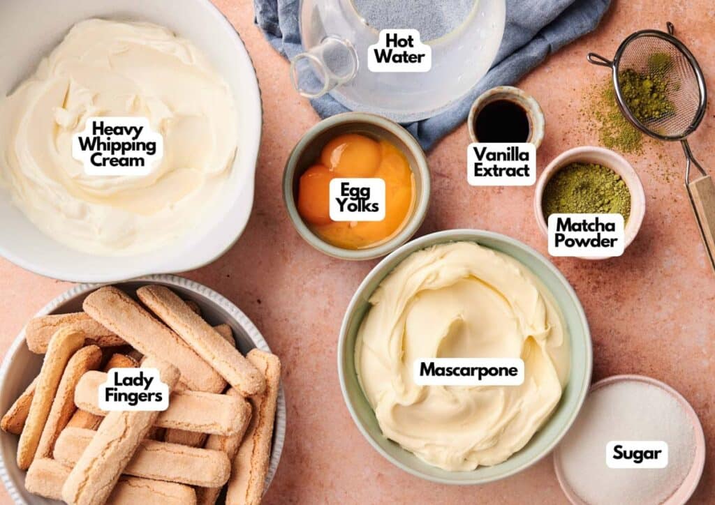 Bowls and dishes with ingredients labeled as heavy whipping cream, egg yolks, mascarpone, ladyfingers, sugar, hot water, vanilla extract, and matcha powder arranged on a countertop for making Matcha Tiramisu.