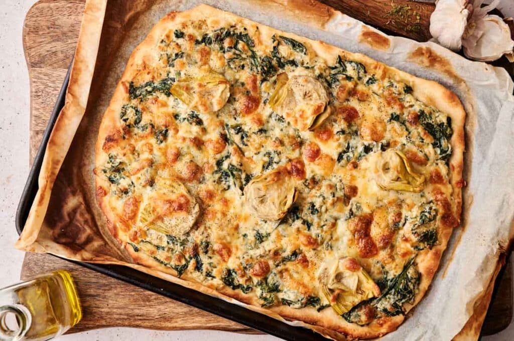 Rectangular flatbread Spinach Pizza topped with artichoke hearts and melted cheese, served on a parchment-lined baking tray, resting on a wooden board.