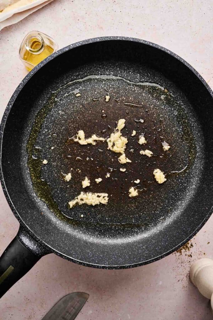 A frying pan with oil and minced garlic sautéing on a stovetop, perfect for preparing toppings for a delicious spinach pizza, surrounded by a bottle of oil and a pepper grinder.
