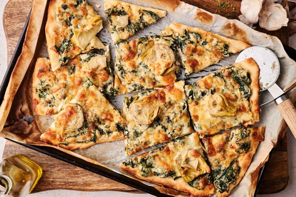 Rectangular spinach pizza with artichoke, cut into slices on a baking tray, surrounded by a pizza cutter, parchment paper, and olive oil.