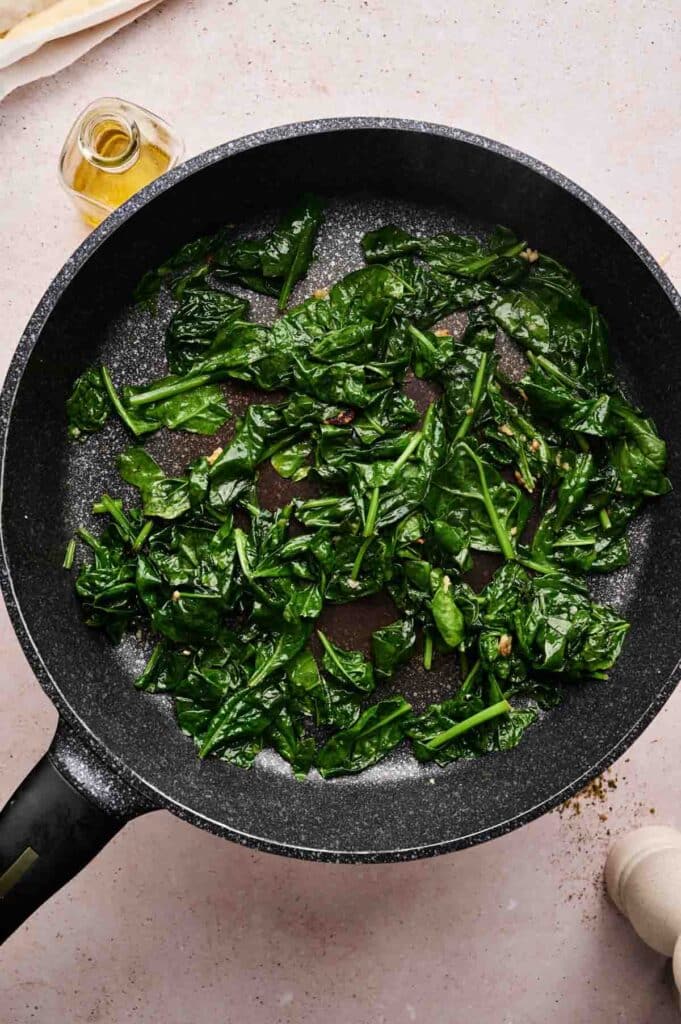 A black frying pan on a light surface contains sautéed spinach leaves, perfect as a topping for spinach pizza. A small bottle of oil and a pepper grinder are visible nearby.