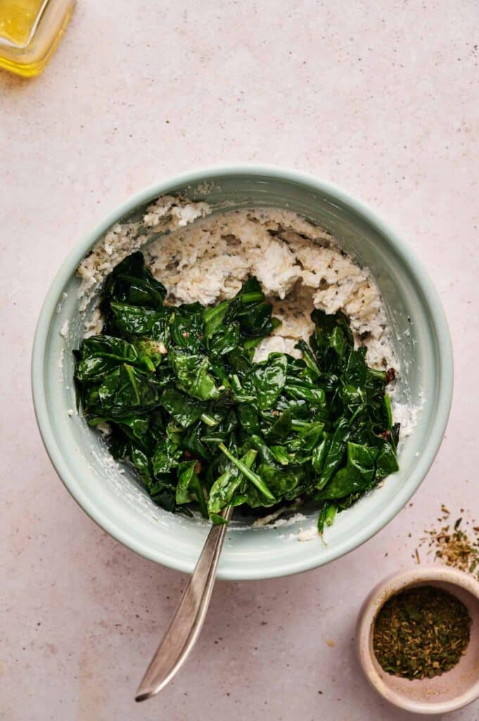 A bowl containing sautéed spinach on top of a white ricotta cheese mixture with a spoon, reminiscent of classic Spinach Pizza flavors, sits next to a small bowl of dried herbs.