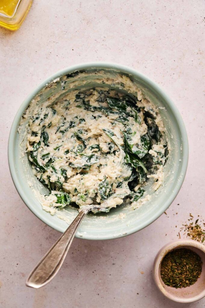 A bowl of creamy spinach and cheese mixture, perfect for topping Spinach Pizza, sits with a spoon on a light surface. A small bowl of dried herbs rests nearby.