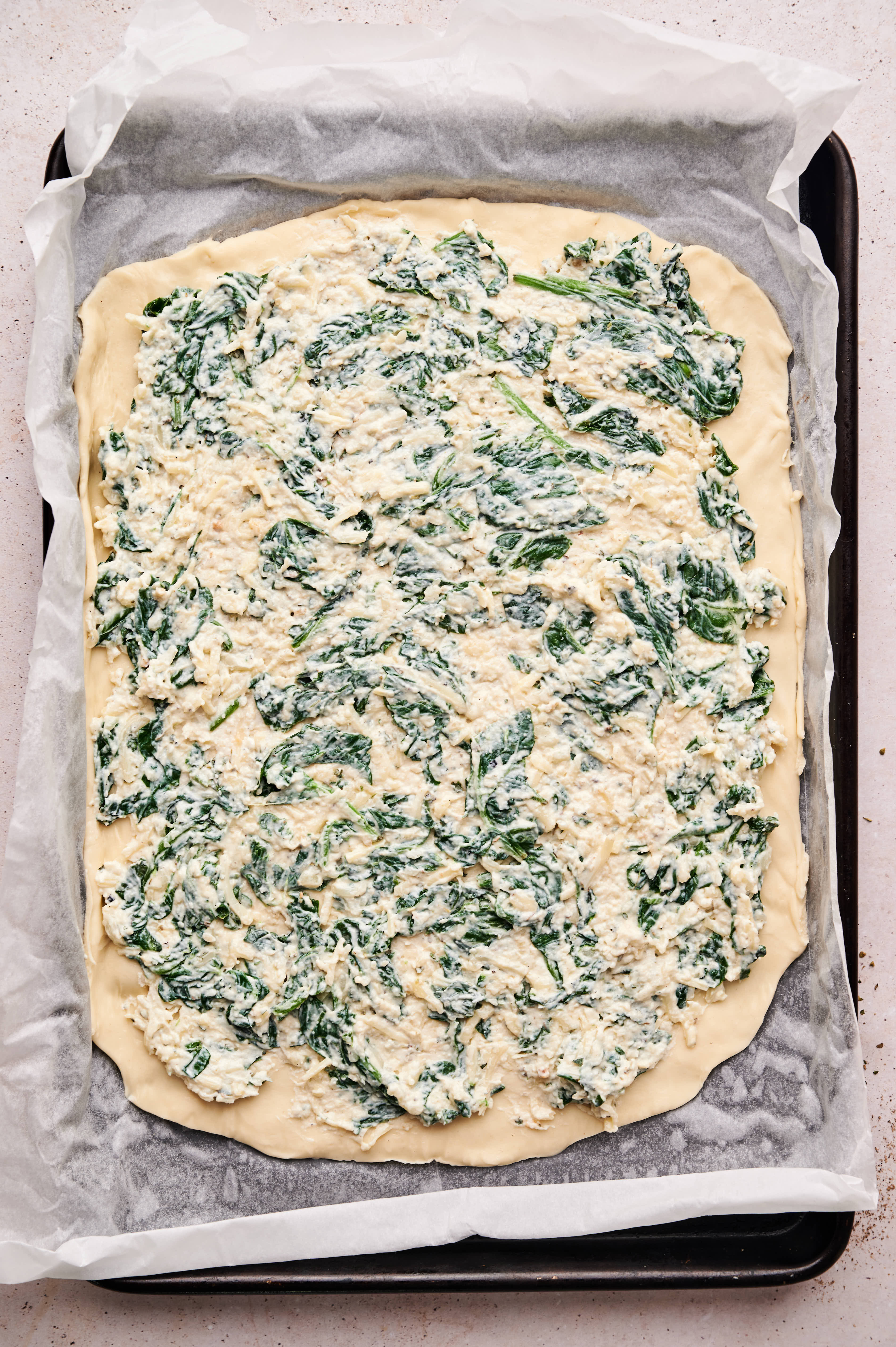 Unbaked rectangular Spinach Pizza dough on a parchment-lined tray, topped with a creamy spinach and cheese mixture.
