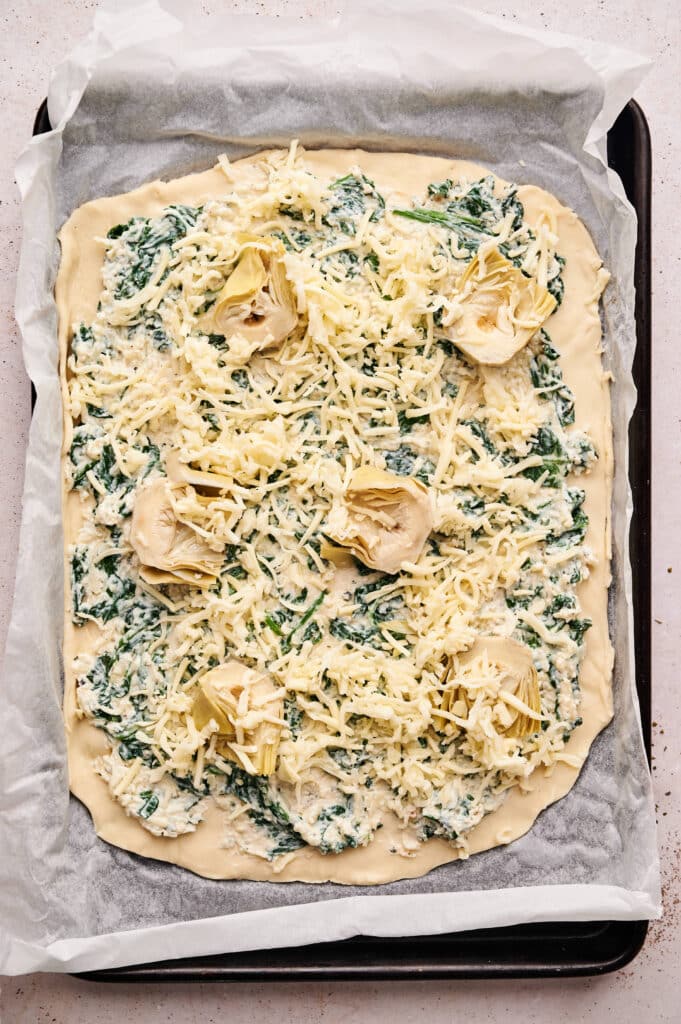 Unbaked rectangular Spinach Pizza topped with cheese and artichoke hearts, arranged on a parchment-lined baking tray.