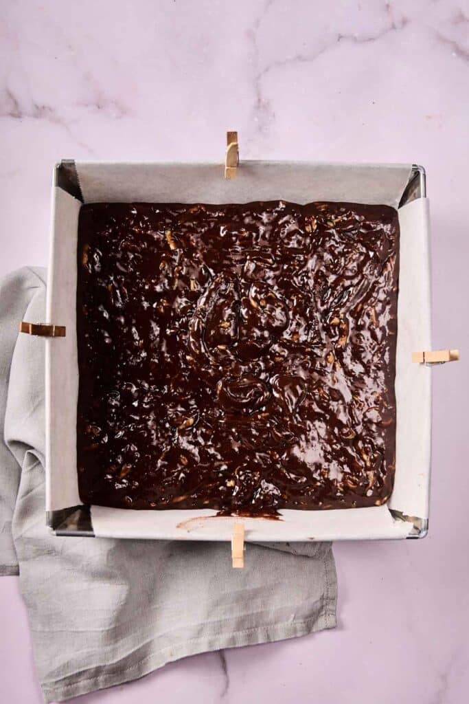 A square baking pan lined with parchment paper and filled with unbaked zucchini brownie batter, secured with wooden clips, sits on a light-colored cloth.