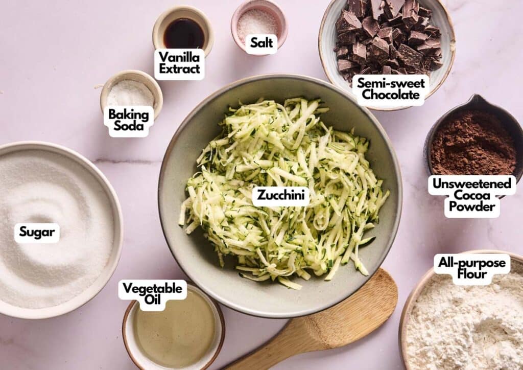 A bowl of shredded zucchini surrounded by labeled baking ingredients: sugar, vegetable oil, vanilla extract, salt, baking soda, semi-sweet chocolate, cocoa powder, and flour—everything you need to make delicious Zucchini Brownies.