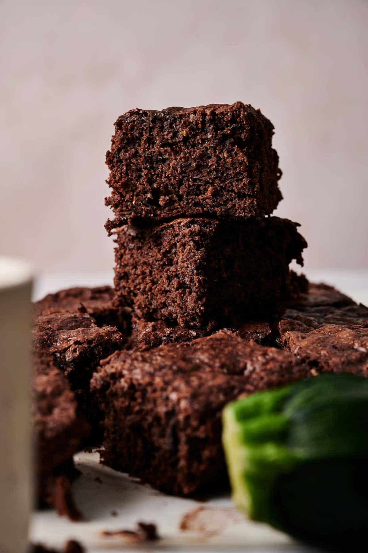 A stack of rich Zucchini Brownies with a visible slice of zucchini in the foreground, placed on a white surface.