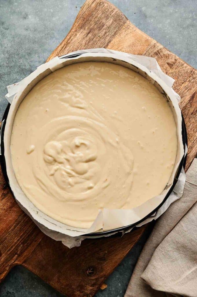 A round baking pan lined with parchment paper is filled with unbaked Cr&egrave;me Br&ucirc;l&eacute;e Cheesecake batter, placed on a wooden cutting board.